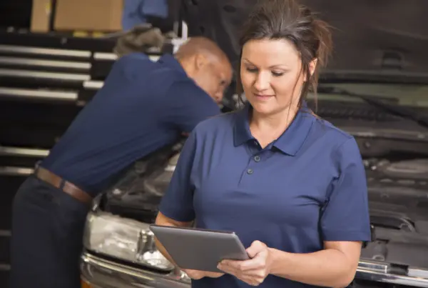 A woman in a navy polo shirt holds a tablet in an auto repair shop while a technician works under the open hood of a car behind her.