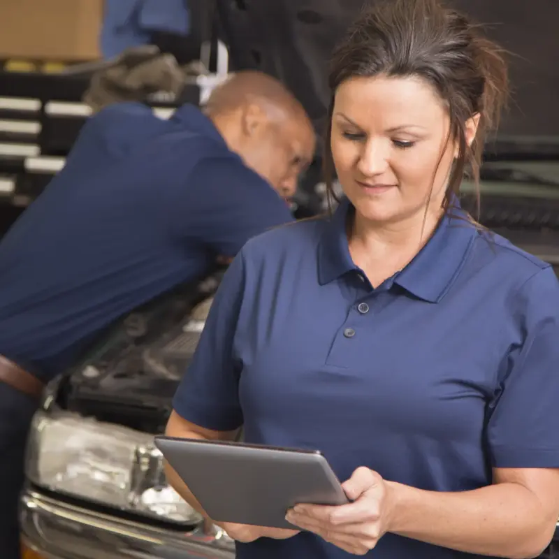 A woman in a navy polo shirt holds a tablet in an auto repair shop while a technician works under the open hood of a car behind her.